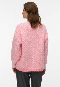 Femme aux cheveux bruns mi-longs portant un pull tricoté rose à motifs, vue de dos, sur un fond blanc uni.