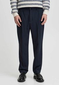 Navy dress pants made of a smooth fabric, featuring a relaxed fit, pleats in front, and an elastic waistband. Black loafers complete the look.