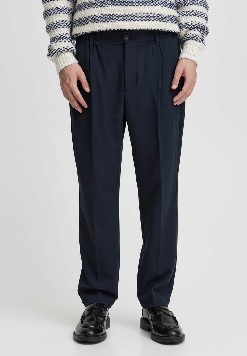 Navy dress pants made of a smooth fabric, featuring a relaxed fit, pleats in front, and an elastic waistband. Black loafers complete the look.