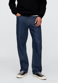 Ciemnoniebieskie jeansy denimowe o prostym kroju nogawek, z klasycznym wzorem pięciu kieszeni i subtelnymi detalami przeszyć. Noszone z czarnymi trampkami.