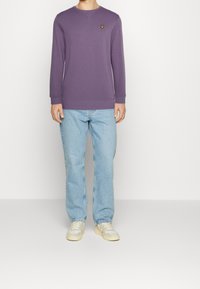 Sudadera morada de algodón con cuello redondo y puños acanalados, que presenta un pequeño logo amarillo bordado. Llevada con jeans de color azul claro.