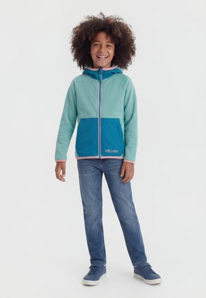 Enfant souriant aux cheveux bouclés, portant une veste à capuche zippée bleu sarcelle et bleu, un jean bleu et des baskets bleues, debout devant un fond uni.