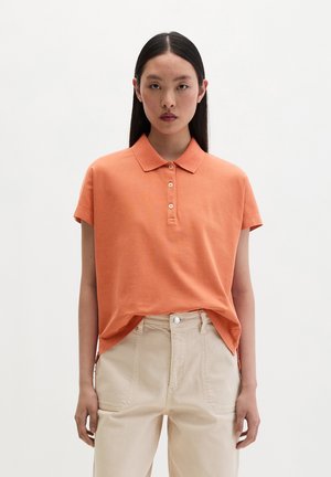 Młoda kobieta z prostymi czarnymi włosami nosi pomarańczową koszulkę polo z krótkim rękawem wpuszczoną w jasnobeżowe spodnie z wysokim stanem, stoi na jednolitym tle.