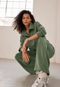 Green pullover tracksuit with a high collar and zip, featuring wide-leg pants. Model wears white sneakers with grey accents. Smooth texture.