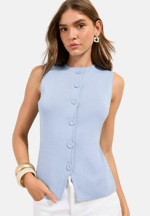 Jonge vrouw die een mouwloze lichtblauwe top met ronde knopen draagt, witte broek, geometrische oorbellen en brede armbanden om haar linkerpols.