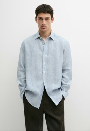 Jeune homme portant une chemise à rayures bleu clair à manches longues boutonnée et un pantalon foncé, debout les mains dans les poches devant un fond uni.