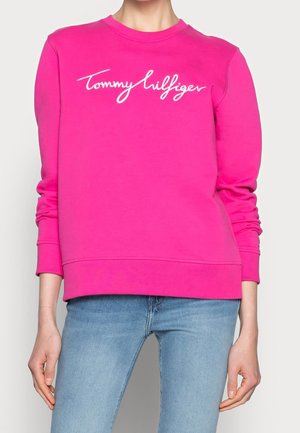 Personne portant un sweat-shirt à manches longues rose vif avec l'inscription blanche "Tommy Hilfiger" sur la poitrine et un jean bleu clair.
