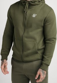 Olijfgroene zip-up hoodie van zacht materiaal, voorzien van een voorzak, verstelbare capuchon en witte logo borduring op de borst.