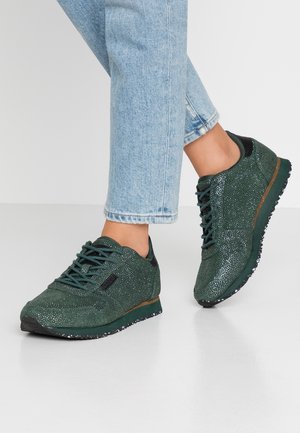 Person iført lyseblå jeans og mørkegrønne glitrende sneakers med sorte såler, stående på en glat grå gulv.