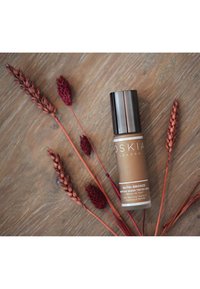 Siero colorato NUTRI-BRONZE in una bottiglia di vetro marrone con un tappo a pompa nero, circondato da rami secchi rossi su una superficie di legno.