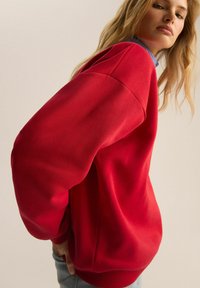 Czerwony sweter z luźnym krojem, szerokimi rękawami i prążkowanym wykończeniem. Stylizowany na niebieską koszulę z kołnierzykiem. Materiał mieszanka bawełny, gładka tekstura.