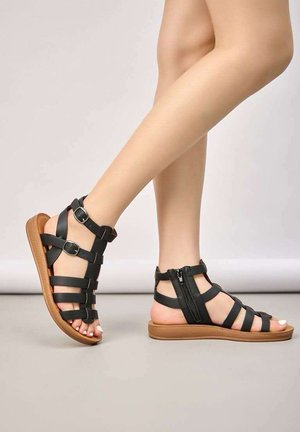 Jambes portant des sandales gladiateur noires avec des boucles et une fermeture éclair sur une semelle marron clair, debout sur une surface lisse et claire.