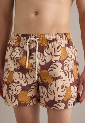 Mann trägt braune Badeshorts mit beigen und gelben tropischen Blattmustern und weißem Kordelbund.