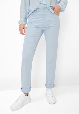 Person trägt hellblaue, aufgekrempelte Hosen, passende hellblaue Bluse und weiße Turnschuhe, steht mit einer Hand in der Tasche.