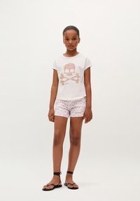 Camiseta de color claro con un gráfico de calavera floral, combinada con pantalones cortos rosas con estampado y sandalias negras de tiras, con lazos en los tobillos.