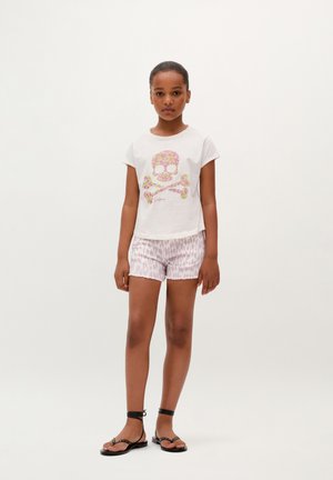 Camiseta de color claro con un gráfico de calavera floral, combinada con pantalones cortos rosas con estampado y sandalias negras de tiras, con lazos en los tobillos.