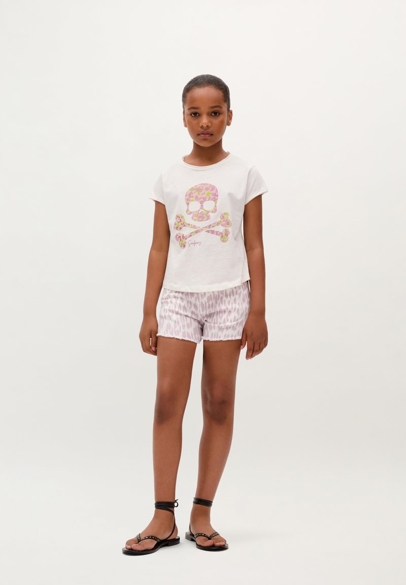 Camiseta de color claro con un gráfico de calavera floral, combinada con pantalones cortos rosas con estampado y sandalias negras de tiras, con lazos en los tobillos.