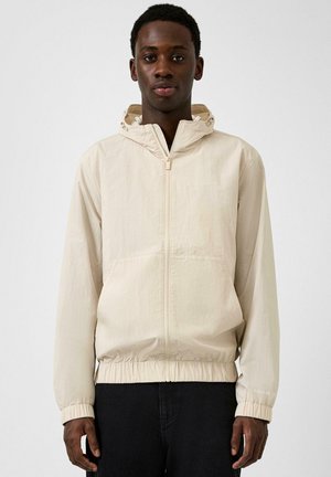 Giovane uomo che indossa una giacca beige chiaro con zip, cappuccio e polsini elastici, in piedi contro uno sfondo bianco uniforme.