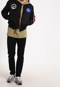 Giacca bomber nera con toppe NASA, chiusura con zip, polsini a costine, indossata sopra un hoodie verde oliva, accompagnata da pantaloni neri e sneaker grigie.
