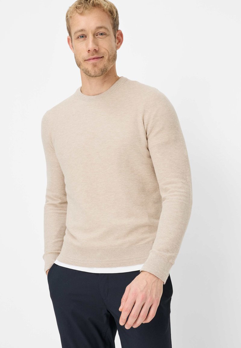 Beiger Pullover mit Rundhalsausschnitt, langen Ärmeln, gerippten Bündchen und Saum. Hergestellt aus strukturiertem Stoff; wird über einem weißen Hemd getragen.