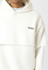 Witte hoodie met een opvallend gelaagd ontwerp. Beschikt over een grote voorzak, zachte textuur en een klein zwart logo op de linkerkant van de borst.