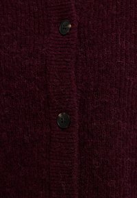 Burgundia värvitud kootud kardigan, millel on tekstuurne muster nähtavate kiududega, mille ees on must nööp.