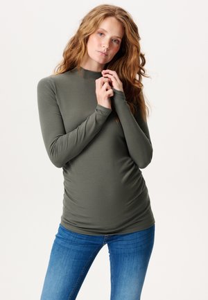 Jeune femme aux longs cheveux roux portant un haut moulant côtelé à manches longues de couleur vert olive et un jean bleu, posant avec les mains près du visage.