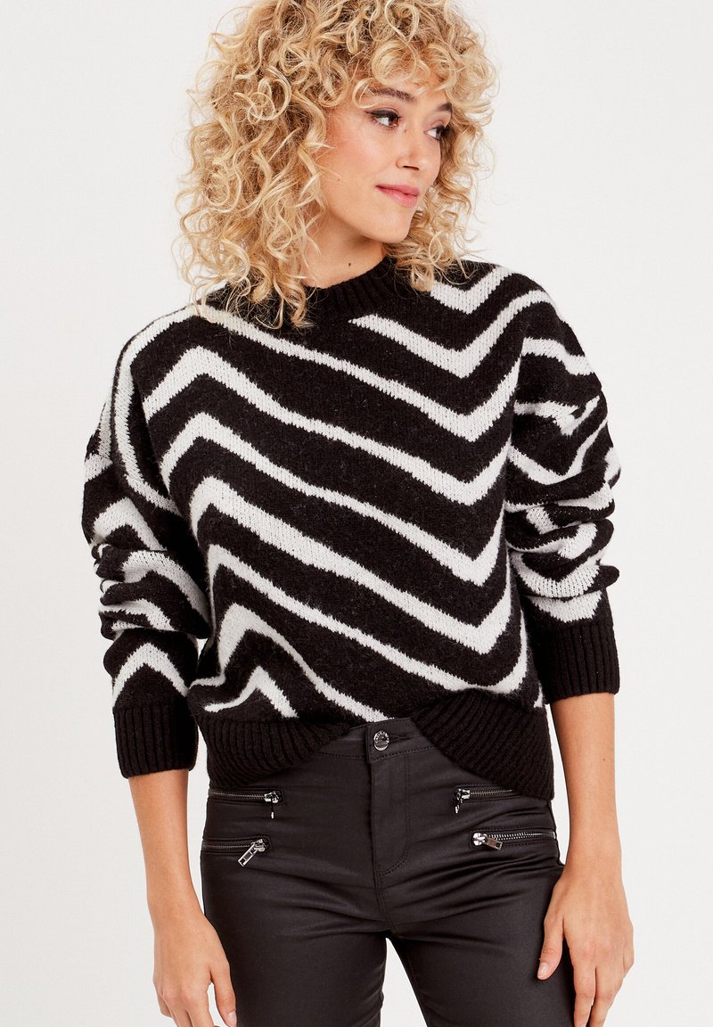 Pull à motif zigzag noir et blanc avec un col rond et des poignets côtelés. Le matériau semble doux et texturé.