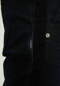 Gros plan sur un jean en denim noir avec une étiquette en tissu visible portant la mention "CHASIN" et un bouton métallique estampillé sur la ceinture.