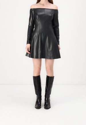 Woman wearing black off-shoulder leather dress with long sleeves and knee-high black textured boots, standing against white background.