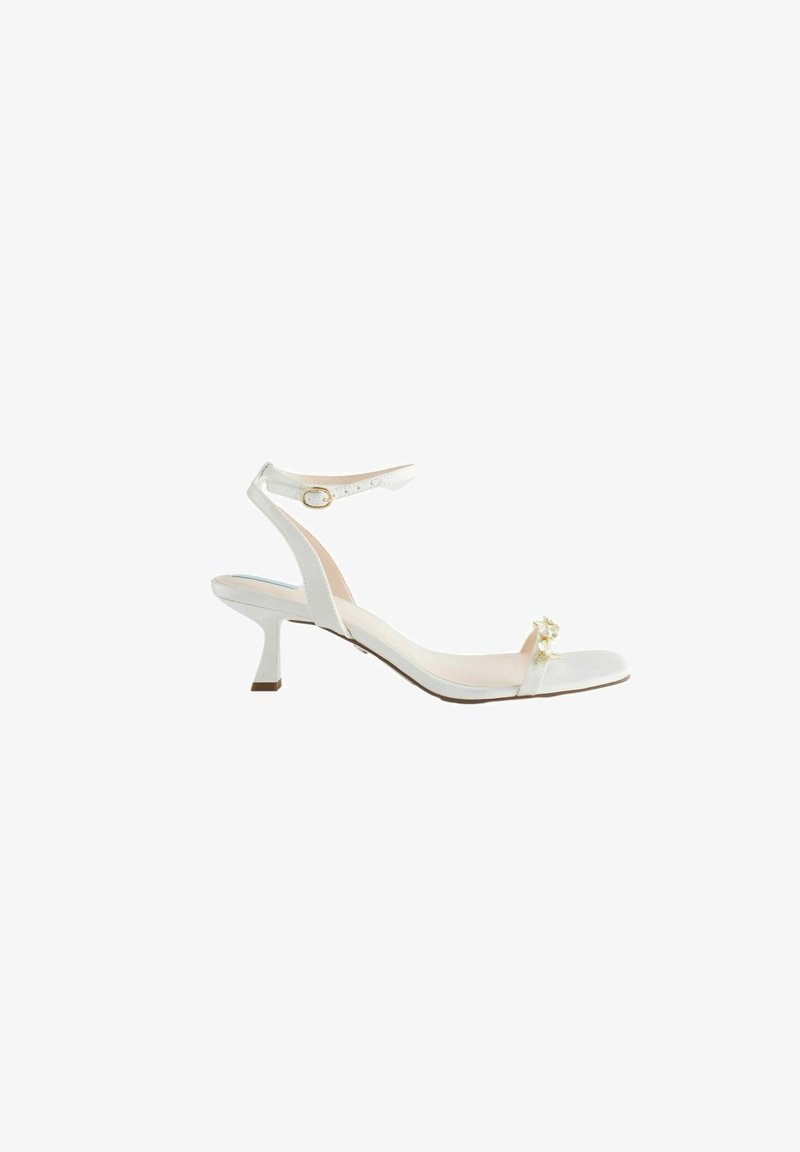 Sandale blanche à talon avec bride à la cheville et boucle dorée, talon bas, bout ouvert et petit ornement floral sur la bride avant.