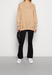 Överstor beige sweatshirt med en vit Nike-logotyp, tillsammans med svarta utsvängda byxor och vita sneakers. Svart quiltad handväska som accent.