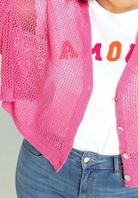 Rosa stickad cardigan med öppen väv, med korta ärmar och silverfärgade knappar. Bärs över en vit skjorta med färgglad text.