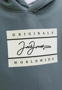 Huvtröja i gråblått, med en vit rektangulär logotyp med svart text "JackJones" och "ORIGINALS WORLDWIDE." Slät tyg.