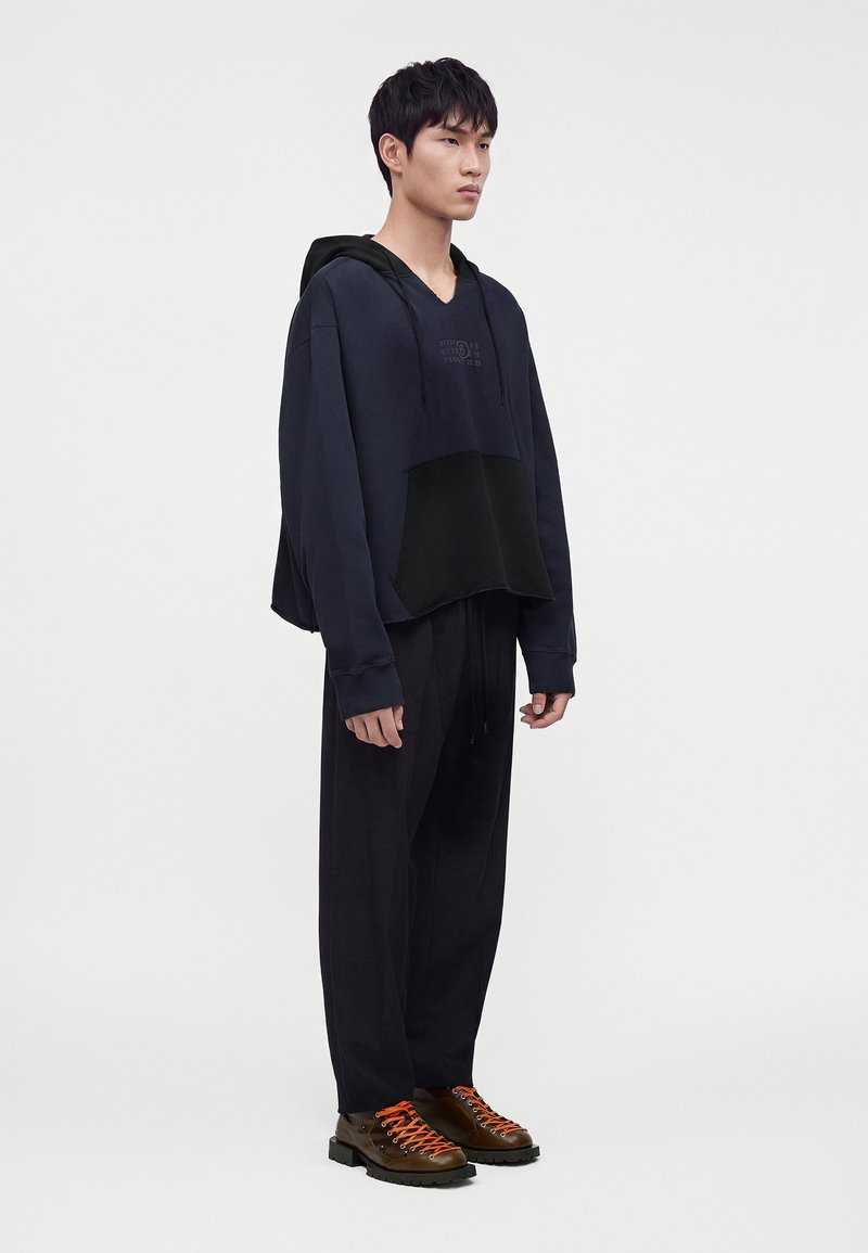 Giovane uomo in piedi che indossa una felpa con cappuccio scura oversize, pantaloni neri larghi e stivali marroni con stringhe di un arancione acceso su uno sfondo chiaro e semplice.