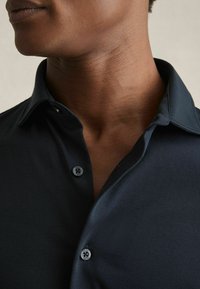 Cămașă button-up de un albastru închis, cu o textură moale și netedă. Are un guler clasic și două butoane negre la gât.