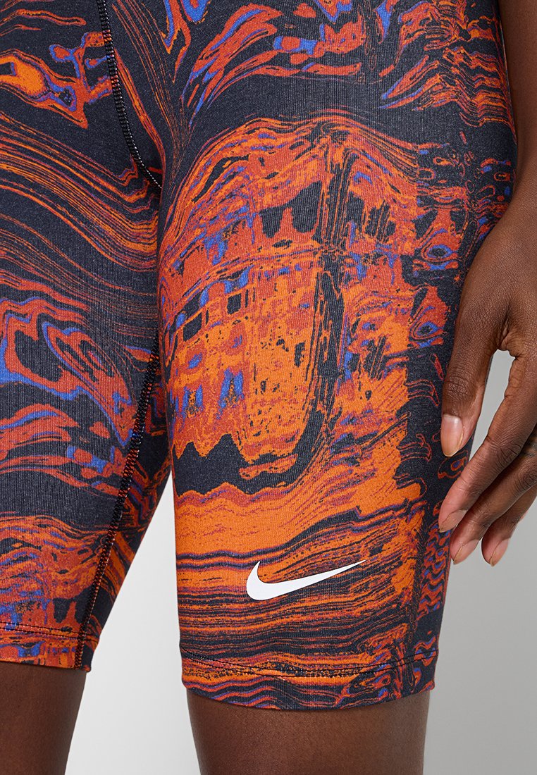Primer plano de una mano de piel oscura descansando sobre el muslo, vestida con pantalones cortos Nike de patrón abstracto en naranja, negro y morado, con el logotipo de swoosh en blanco.