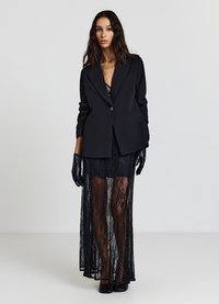 Schwarzer Blazer mit tailliertem Schnitt, mit Ein-Knopf-Verschluss, getragen über einem transparenten schwarzen Spitzenkleid. Lange Ärmel und passende Handschuhe verleihen Eleganz.