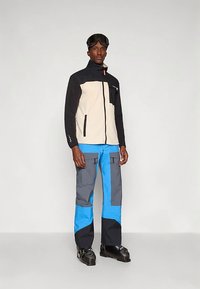 Giacca in pile crema e nera con chiusura zip, abbinata a pantaloni impermeabili grigi e blu con tasche cargo e una vestibilità rilassata.