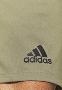 Harmaat urheilushortsit, jotka on valmistettu sileästä ja joustavasta kankaasta, sisältävät näkyvän mustan Adidas-logon vasemmassa alareunassa.