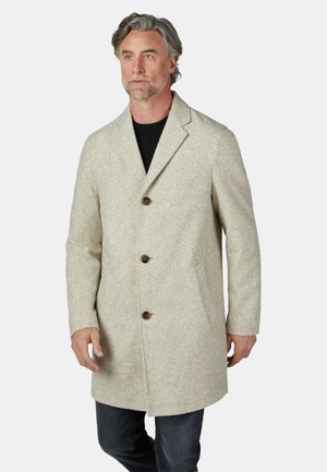 Mann mit grauem Haar, der einen hellbeigen, geknöpften Wollmantel über einem schwarzen Hemd und dunklen Hosen vor einem schlichten hellen Hintergrund trägt.