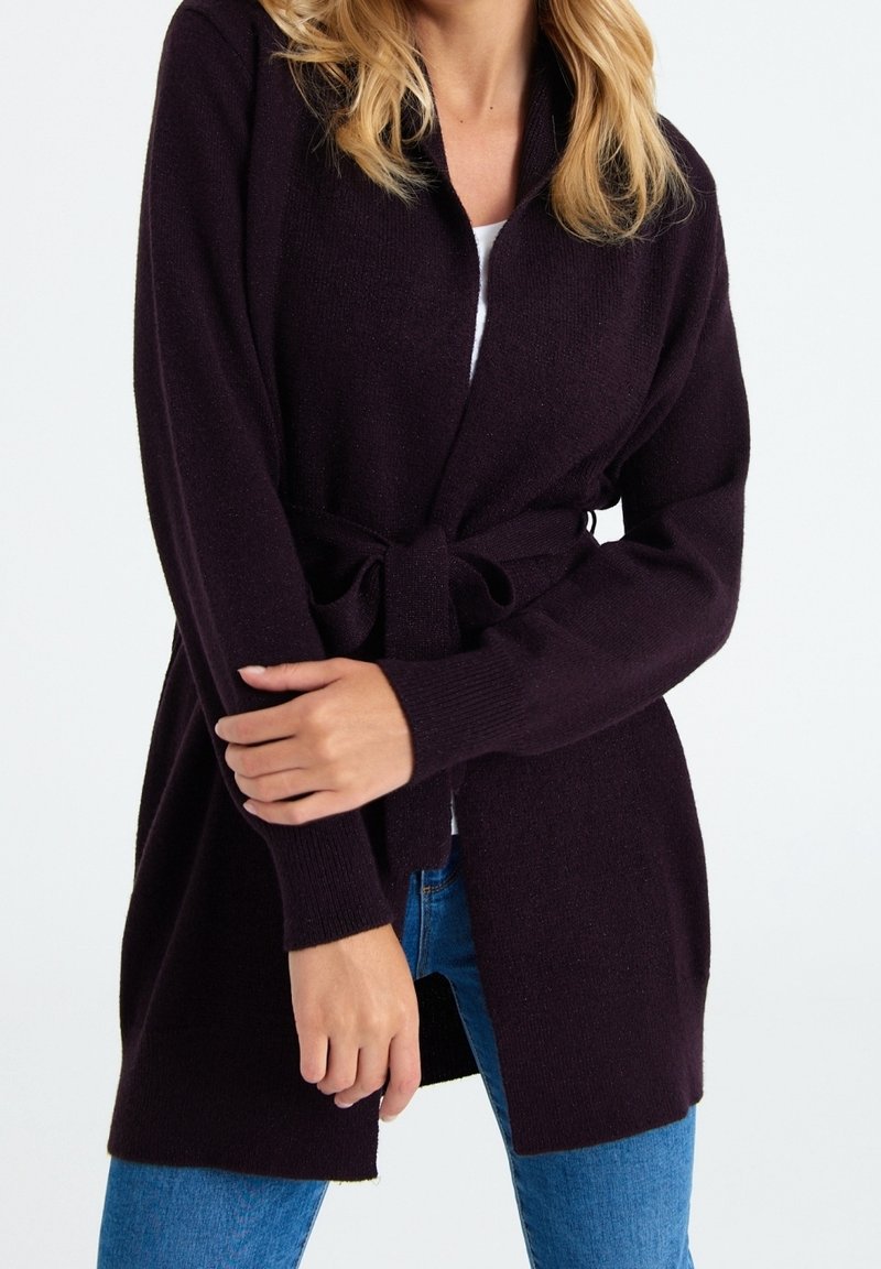 Femme portant un cardigan violet foncé ceinturé sur un haut blanc et un jean bleu, debout les bras croisés devant.