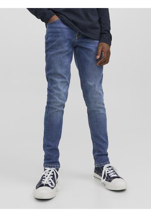 Person iført slim-fit blå jeans, navy sneakers med hvide såler og snørebånd samt en mørk langærmet skjorte, stående på en neutral baggrund.