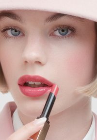 Kusbestendige lippenstift in een slanke zwarte en gouden tube, aangebracht op volle lippen. De tint is een zachte koraalroze met een glanzende afwerking.