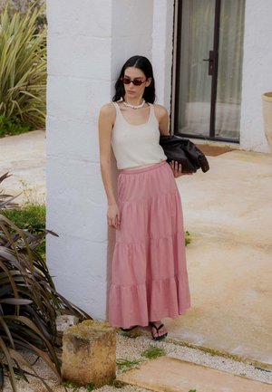 Frau mit Sonnenbrille, weißem Tanktop, pinkem gestuftem Rock und schwarzen Sandalen lehnt draußen an einer weißen Wand und hält eine schwarze Jacke.