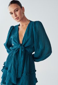 Tirkizna haljina s teksturom, dugim napučenim rukavima, dubokim V-izrezom i pojasom s vezanjem. Ima slojevite čipke na rubu i mekani, lagani materijal.