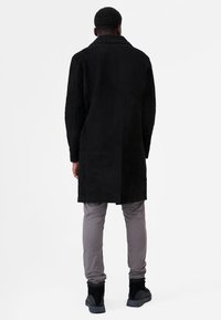 Sort ruskind frakke med høj krave, der har en glat tekstur og struktureret design. Sammen med lysegrå bukser og sorte støvler.