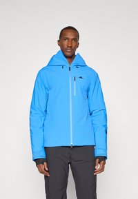 Casaco impermeável azul com capuz, fecho frontal, logotipo em preto e punhos texturizados, usado sobre calças escuras.