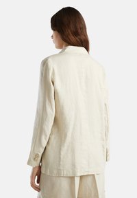 Beżowa lniana marynarka z wyciętym kołnierzem, długimi rękawami z zapięciem na guziki oraz luźnym krojem, prezentująca fakturowany materiał.