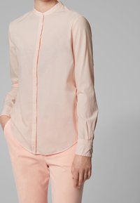 Chemise rose clair à manches longues avec un col montant, devant boutonné et texture légèrement côtelée. Ourlet arrondi et poignets ajustés. Associée à un pantalon rose clair.